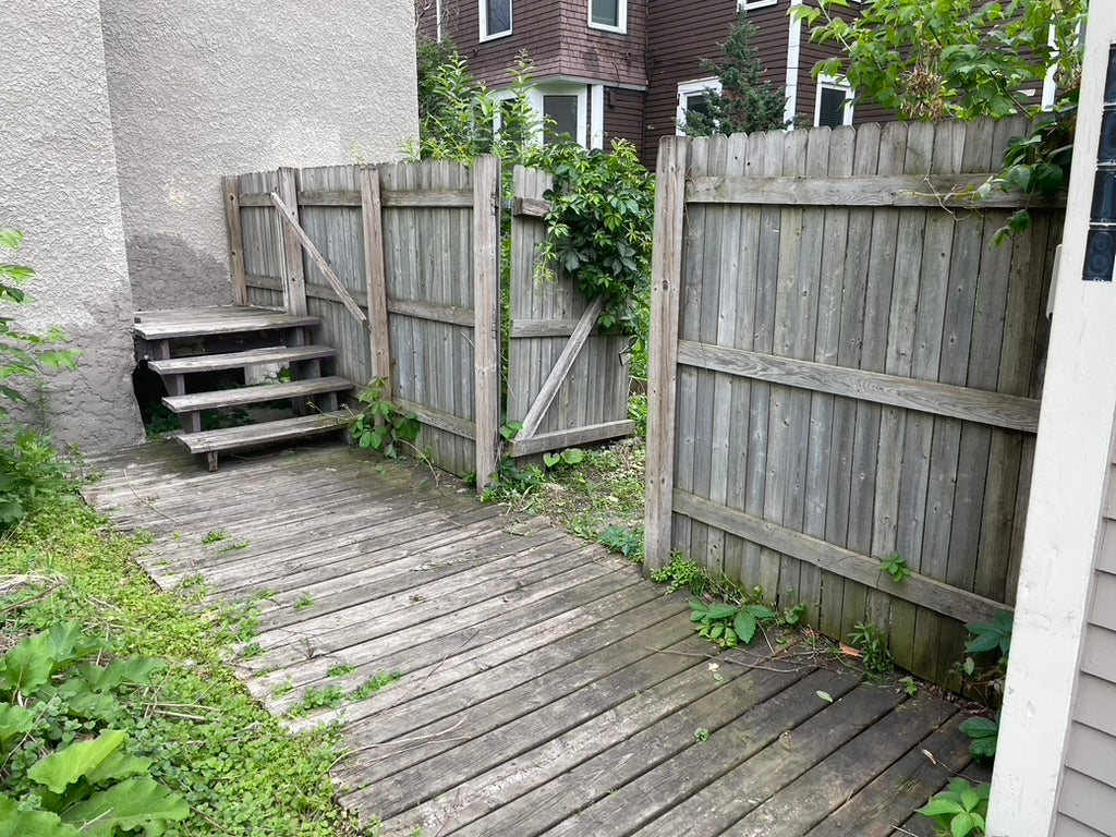 Before urban backyard renovation in Minneapolis showing old wood platform deck, uneven ground, and overgrown planting near fence line.